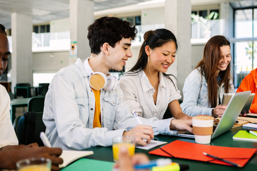 Group of happy international students studying together on laptop computer sitting on cafeteria table at college campus